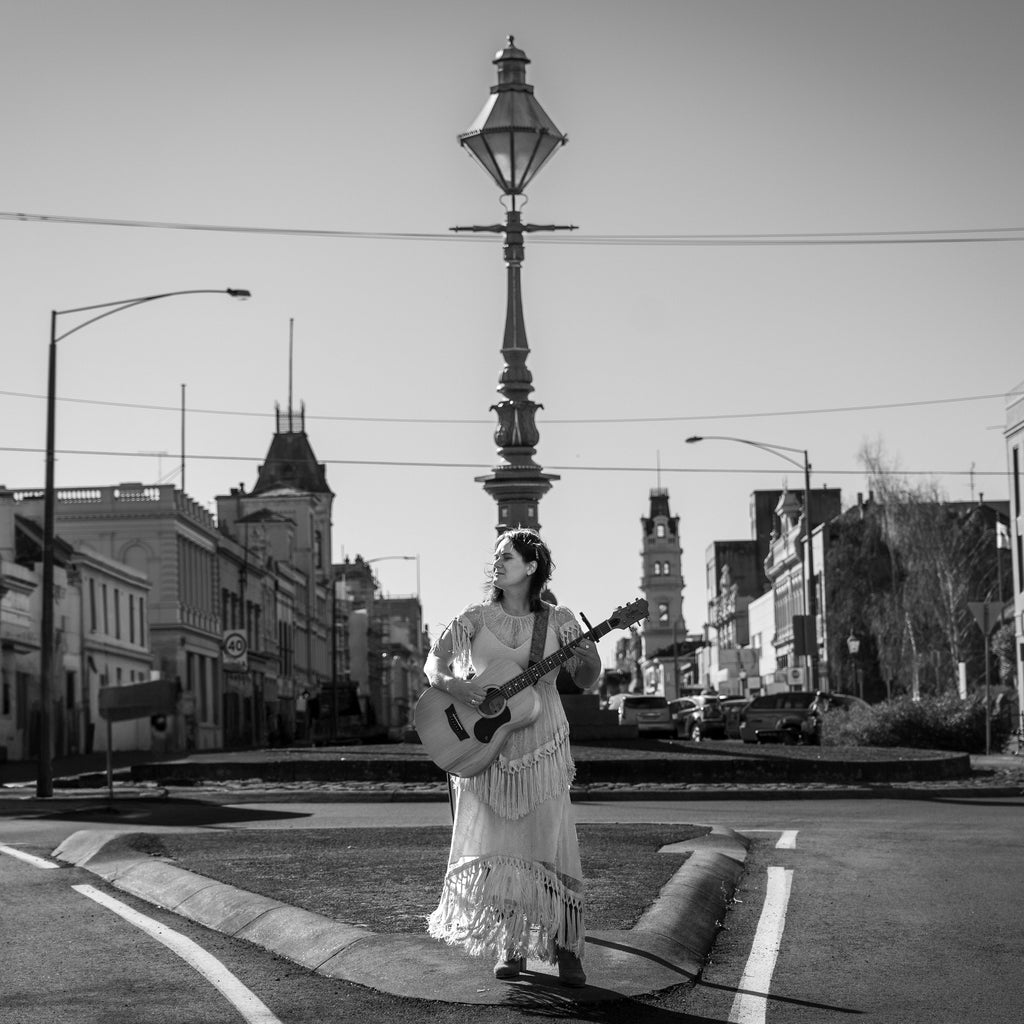 Person in a long dress holding a guitar on a city street with buildings and streetlights in the background.